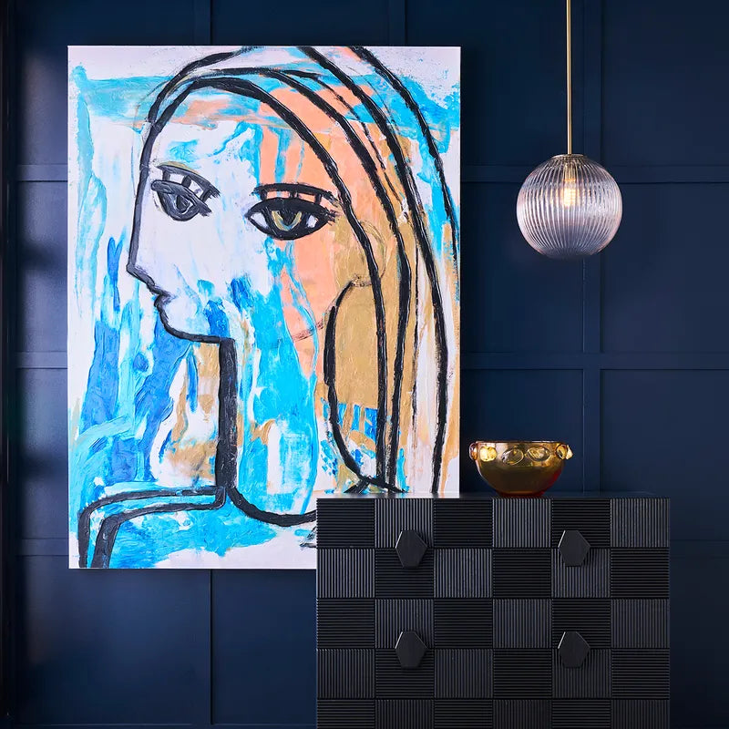Modern interior with navy panelled wall featuring a bold abstract portrait painting in blue, peach, and white tones, black geometric cabinet, gold decorative bowl, and glass pendant light